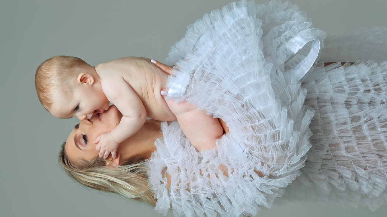 Mother holds and kisses her beloved naked baby. Happy motherhood concept. White backdrop. Vertical screen.