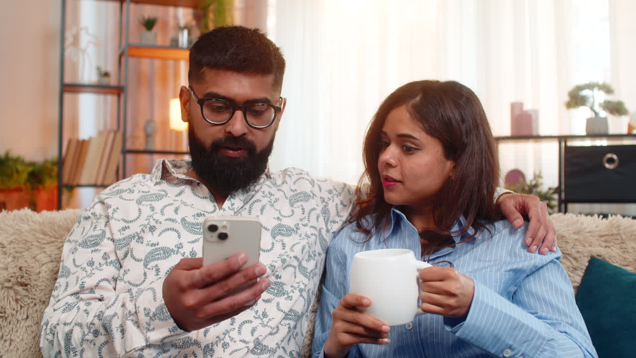 Indian family couple man woman drinking morning coffee using smartphone together calm start of day