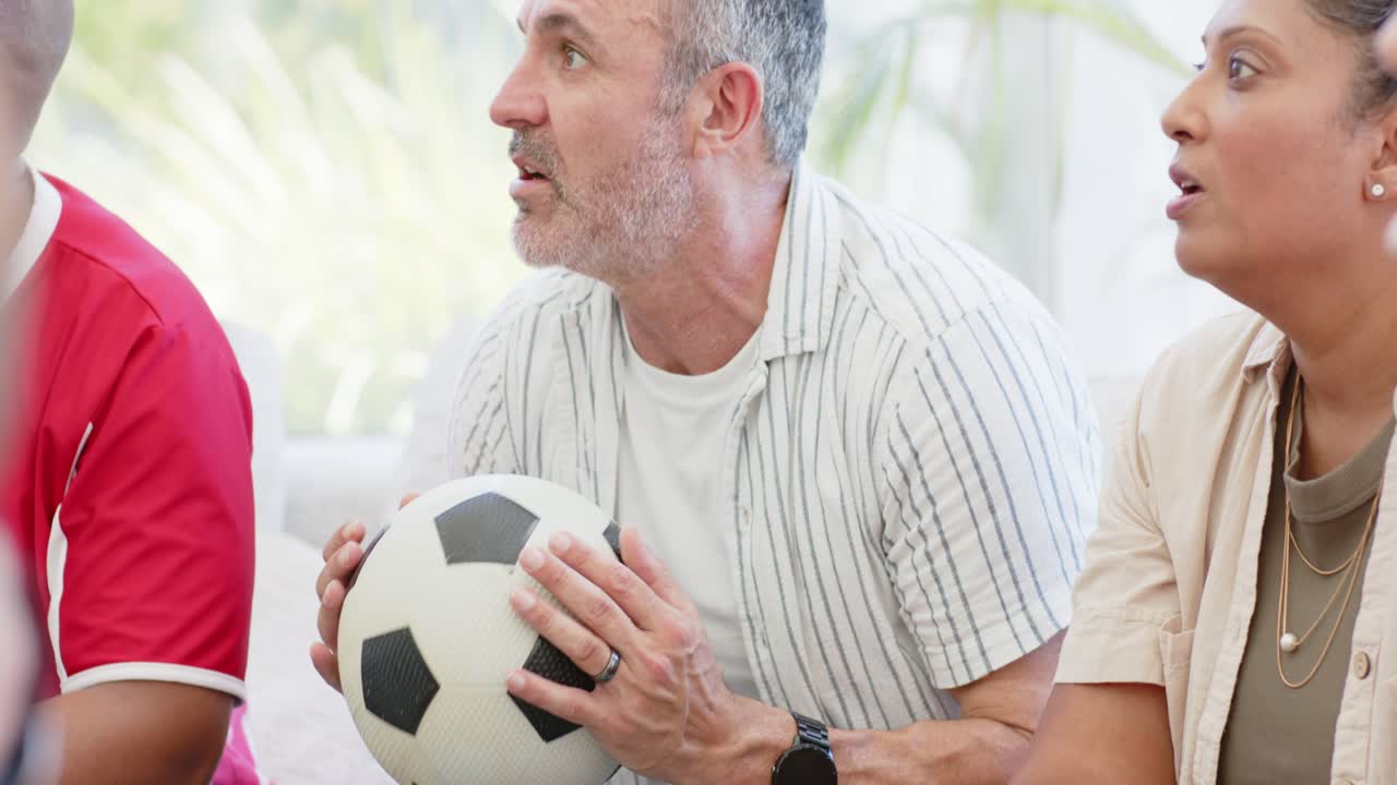 Amici di diverse etnie che guardano una partita di calcio sul divano, con l'uomo al centro che tiene un pallone bianco e nero e applaude.