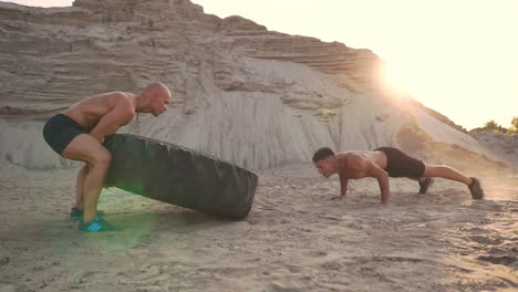 Two-athletes-train-in-active-mode-on-the-beach-doing-push-UPS-and-pushing-a-huge-wheel