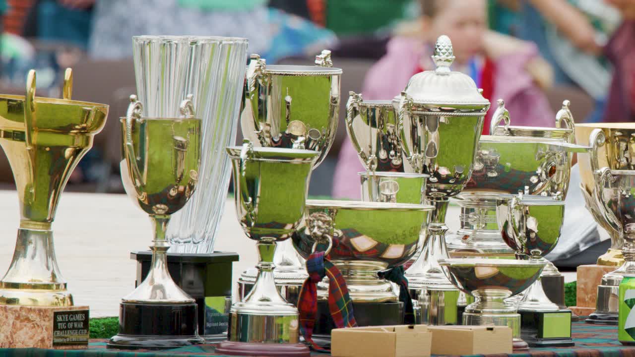 Silver and gold trophies displayed outdoors on table, with blurred spectators in daylight background