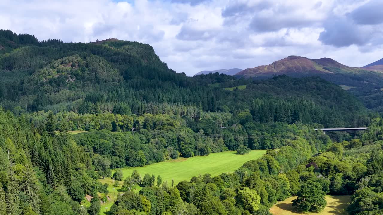 Drone camera smoothly pans across green forest, river valley, and distant mountains under daylight