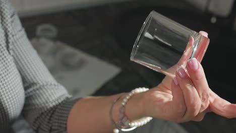 close up of transparent glass cup carefully observed by woman with pink polished nails and bracelet against soft kitchen background, showcasing clean glass inspection under bright natural light