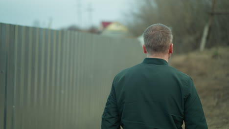 a close view of a man walking along a fence, captured from behind, with a view of back of his head and jacket, with a blurred background of an industrial area