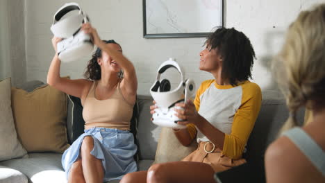 Diverse female friends laughing and exploring virtual reality headsets on cozy sofa, at home