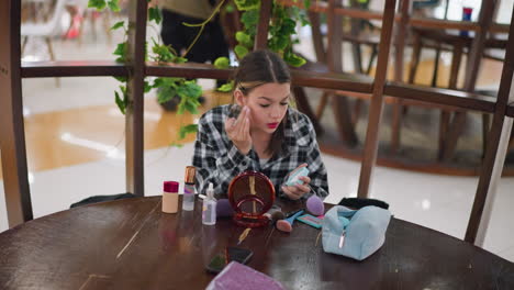 joven aplicando polvo en su cara usando la mano, con productos de maquillaje como fundación, rubor y pinceles esparcidos en la mesa frente a ella, el espejo y las herramientas de belleza son visibles en la escena