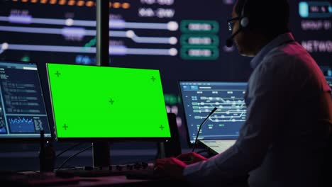 professional it technical support specialist and software developer working on computer with green screen mock up display in monitoring control room with digital screens. employee puts on headphones.