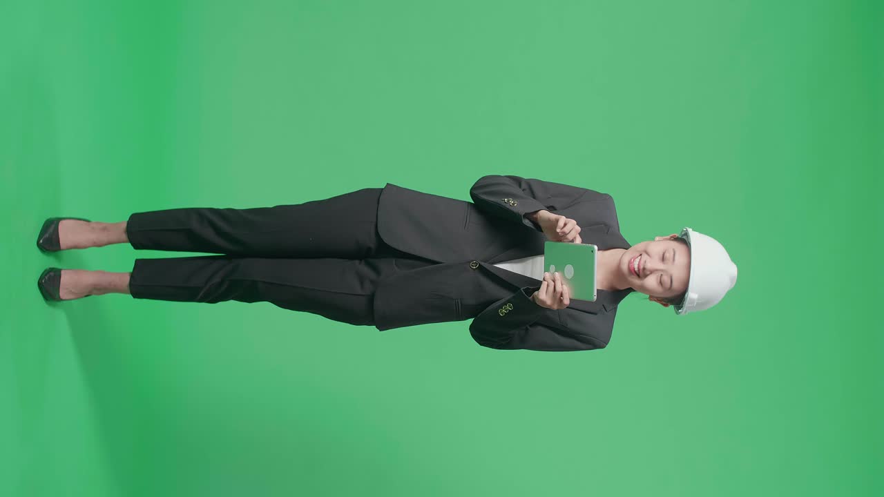Full Body Of Smiling Asian Female Engineer Looking Around And Shaking Her Head While Using Tablet In The Green Screen Studio