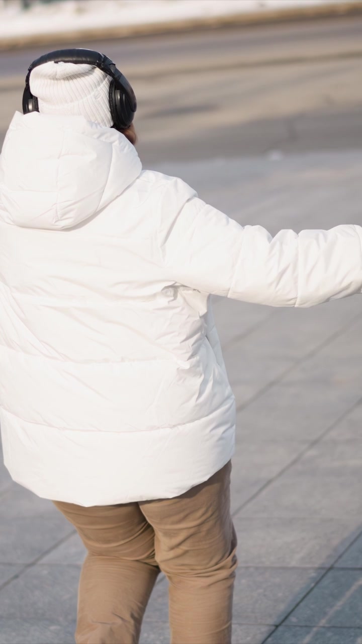 Woman wearing white puffer jacket and black headphones walks along snowy sidewalk occasionally looking down at her hands. scene features urban surroundings with snow piles and street