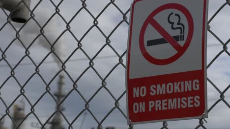 a refinery pipe smoking and polluting blurred in the background with a no smoking sign in the foreground on a chain-linked fence