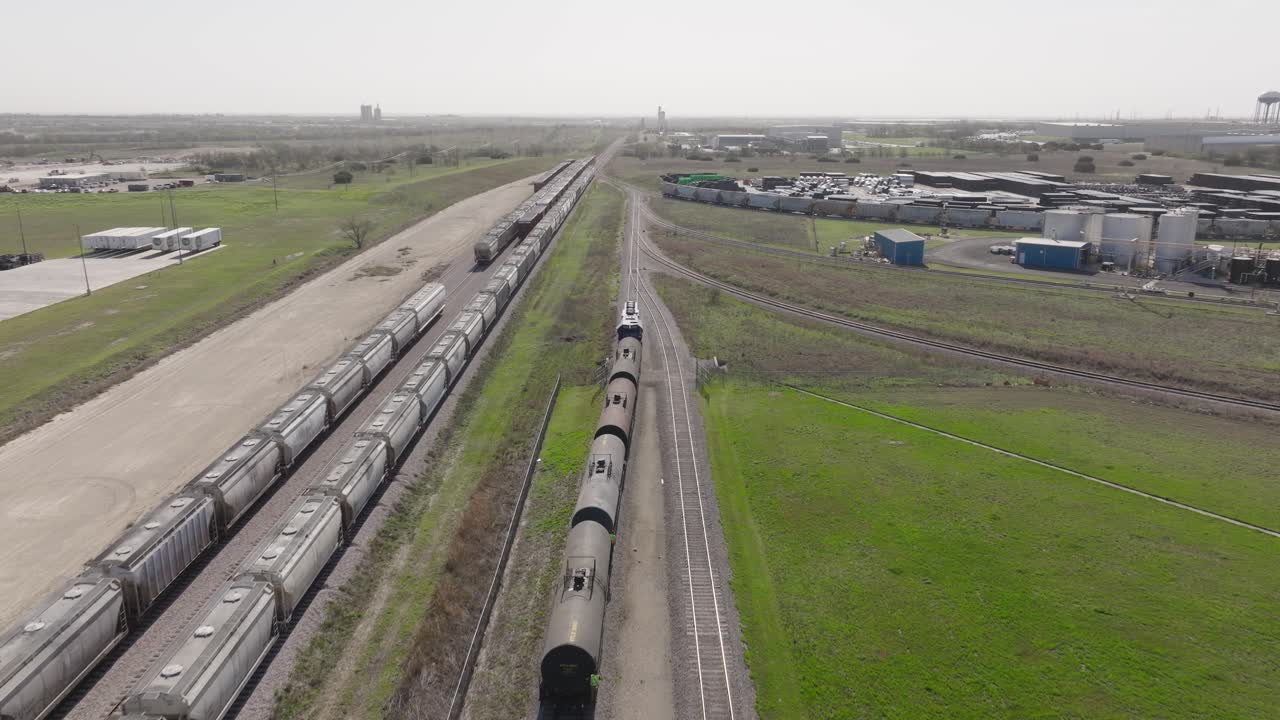 Aerial tracking shot of an oil train (tank cars) pulling out of an industrial rail yard past freight cars on a sunny day, showcasing energy logistics