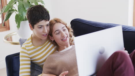 una feliz pareja de lesbianas caucásicas acostadas en el sofá, usando una computadora portátil y sonriendo en una casa soleada