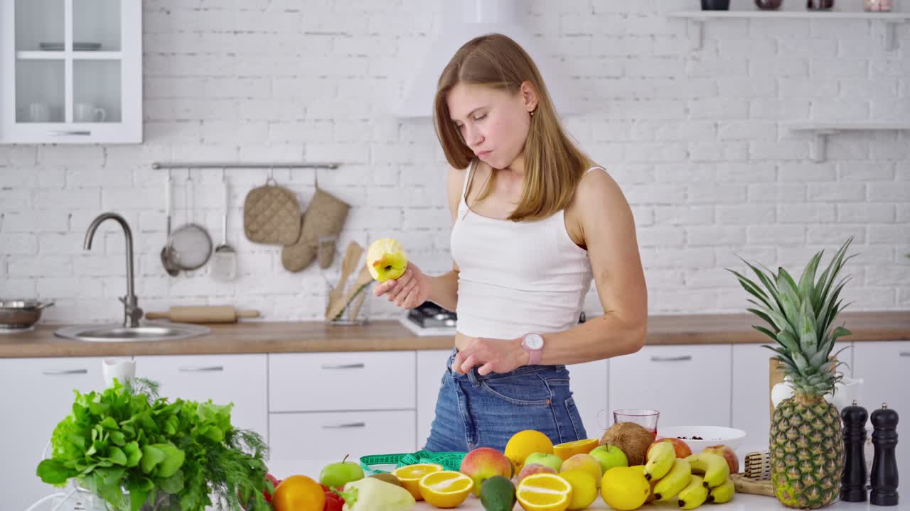 Losing weight concept. Young woman eating fruit in the kitchen. Beautiful model in jeans standing at the table full of organic vitamins.