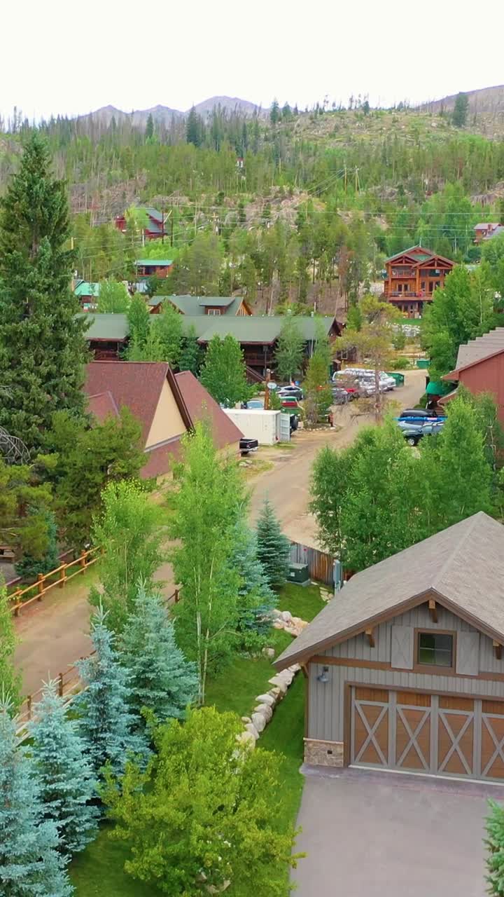 Un drone video verticale si alza tra gli alberi sempreverdi per rivelare una tranquilla città di montagna con baite, strade tortuose e pendii boscosi, aprendosi su un pittoresco quartiere alpino in una valle aspra