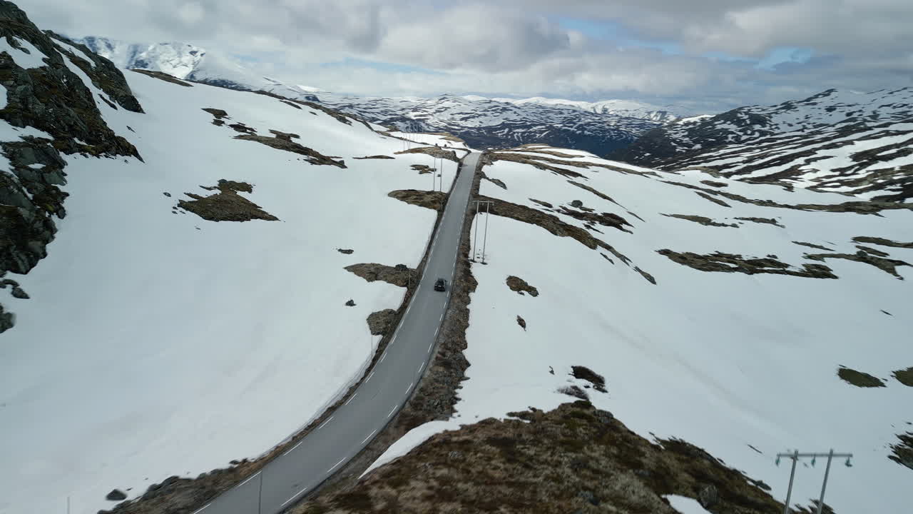 Drone shot of a car on the Sognefjellet tourist road in Norway. Large snowfields pile up on either side of the road. As the camera pans, the mountain landscape in the background becomes visible