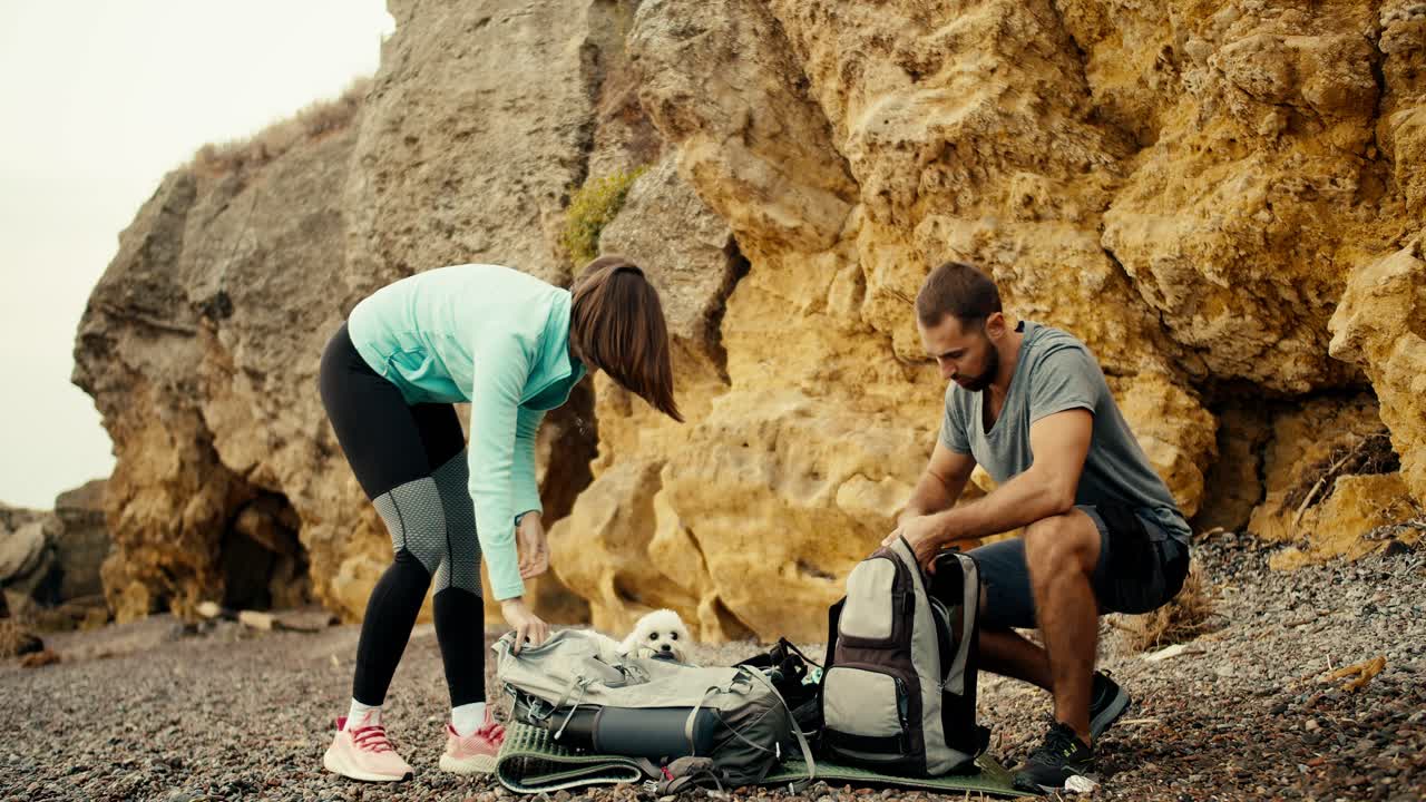 una chica rubia con una chaqueta azul y un tipo moreno con una camiseta gris se acuestan y sacan municiones de sus mochilas en una playa rocosa cerca de las rocas amarillas, estarán escalando rocas durante el día