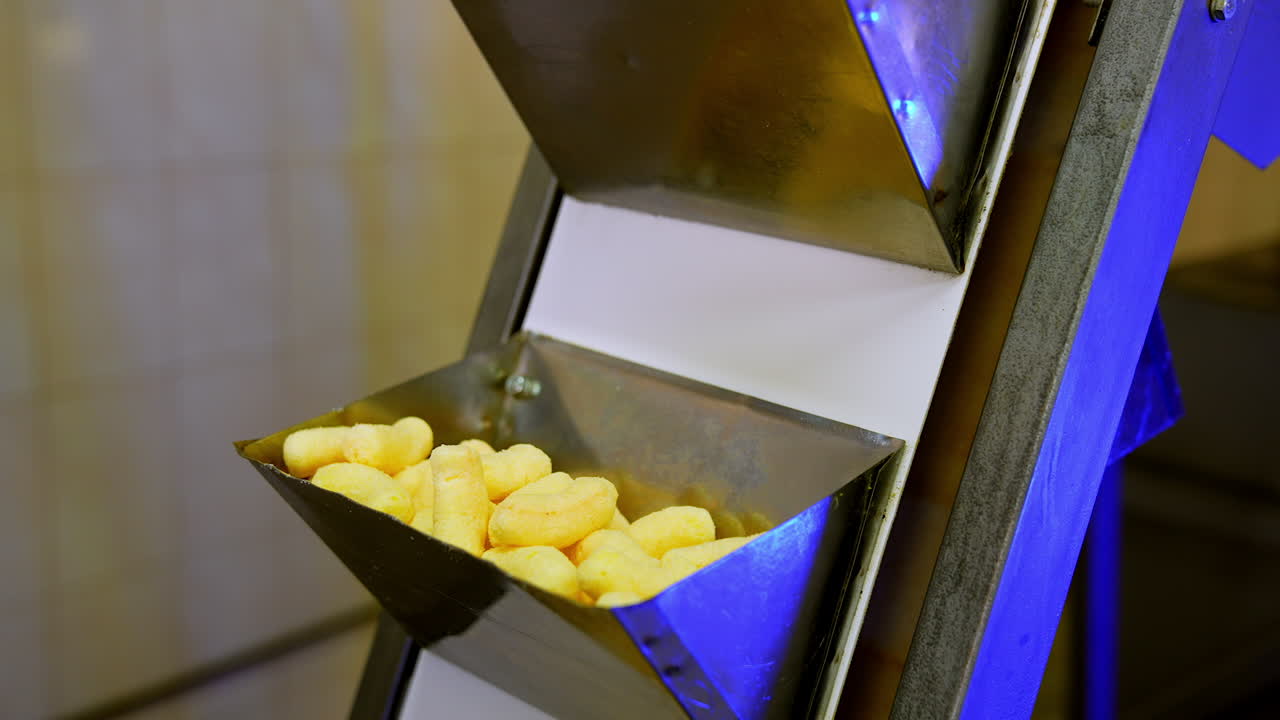 Metal pockets at conveyor belt filled with sweet corn sticks move up. Snack desserts manufacturing close up.