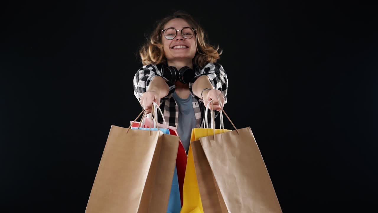 alegre hermosa joven europea con gafas y auriculares en el cuello sosteniendo coloridas bolsas de compras en la temporada de venta, felizmente mostrarlos en las manos extendidas aislados en fondo negro