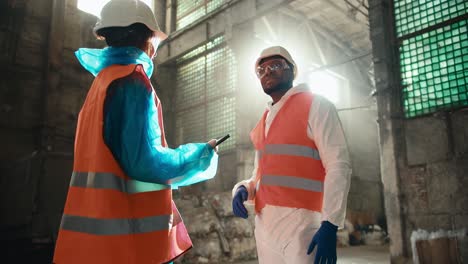 Two-colleagues-in-special-uniforms-and-orange-vests-communicate-at-a-large-gray-waste-recycling-plant.-A-man-and-a-girl-communicate-at-a-waste-processing-plant