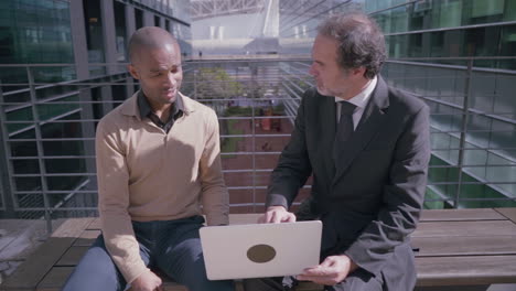 Adult men discussing business projeсt with laptop and shaking hands outdoors