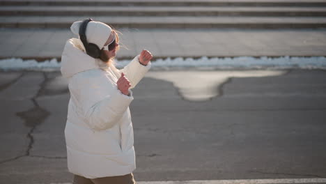 young artist walking down tiled street wearing headphones and beanie, moving to music beat against snowy curb and urban stair backdrop under winter sunlight, expressive arms and joyful smile
