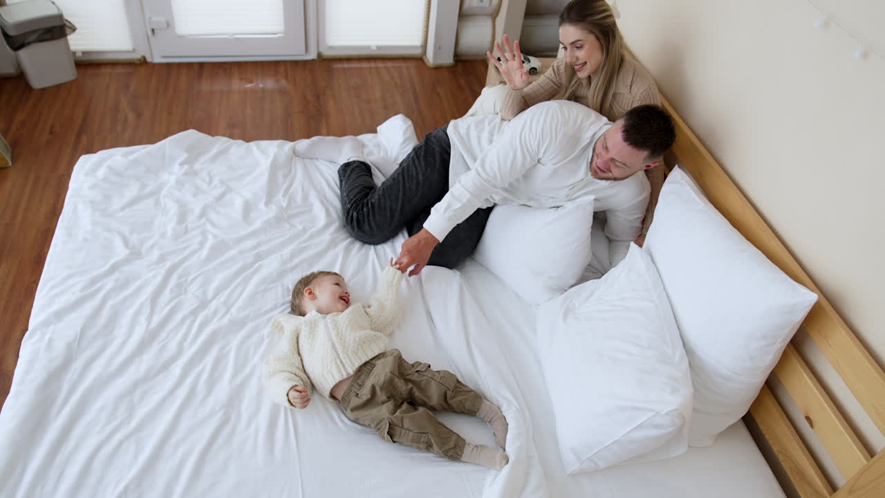 Family of three sits on the big double bed. Dad tickles a little baby boy and kid laughs adorably. Top view.