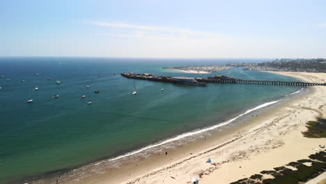 Toma-Aérea-Volando-Sobre-Una-Playa-De-Arena-De-California-Con-El-Muelle-Stearns-Wharf-Y-El-Puerto-Azul-Claro-Del-Océano-Lleno-De-Veleros