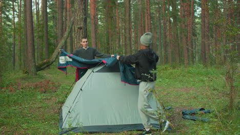 young campers carefully spread and adjust tent cover over pitched tent in forest clearing, working together with focus and coordination surrounded by green trees and natural outdoor environment