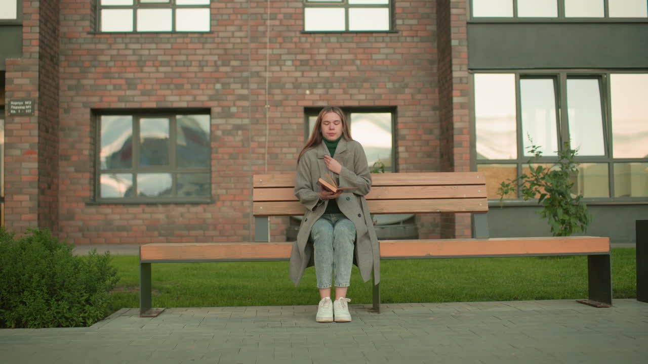 Student seated on wooden bench outdoors reading book while adjusting coat, wearing gray overcoat and green turtleneck, studying thoughtfully with brick residential building background