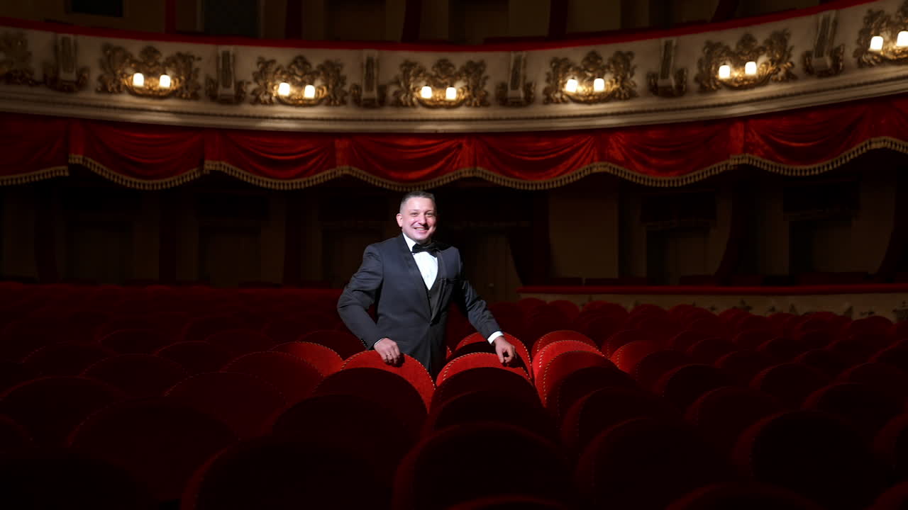 Man stands alone among velevet chair in classic theater. Male in classic suit invites public come in with gesture. Time before performance.
