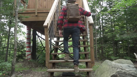 a young man climbs up the stairs to an elevated small cabin while wearing flannel and jeans with a leather back in slow motion, 120 fps