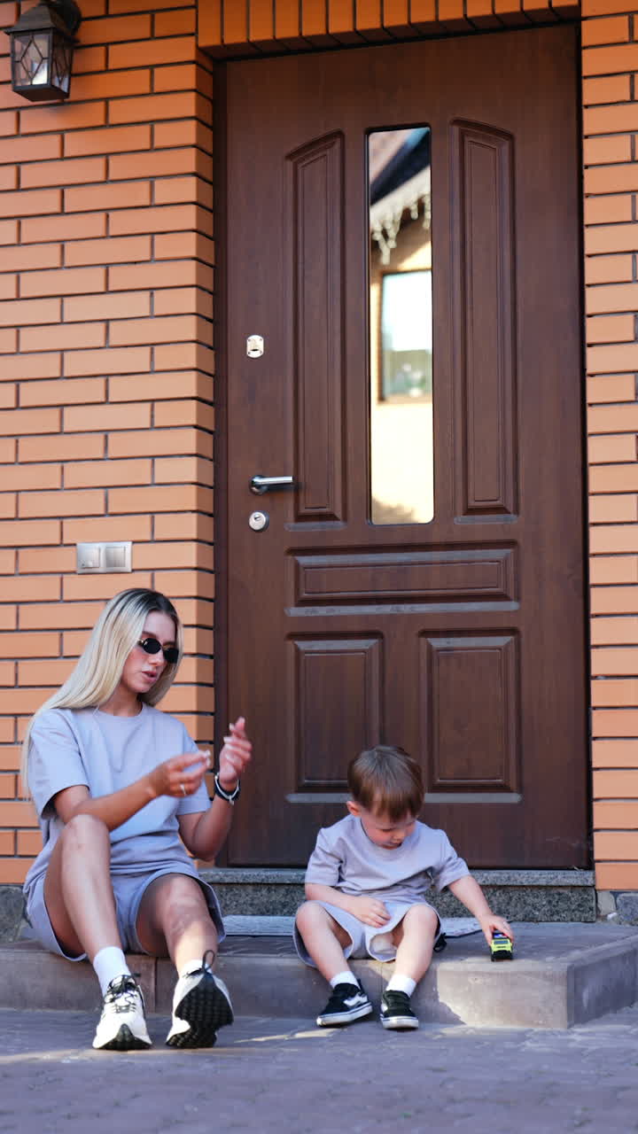 Young Caucasian woman with long blonde hair sits on the doorstep with her little baby. Lovely child plays with a toy car. Vertical video