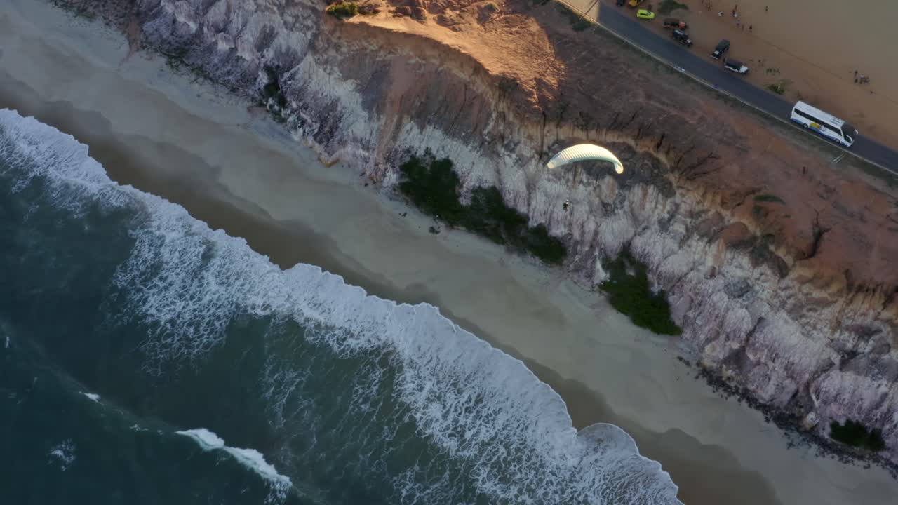 Aerial drone bird's eye top view rotating and following a paraglider flying over the Cliffs of Cacimbinhas in Pipa, Brazil Rio Grande do Norte with golden sand dunes, exotic green foliage, and a beach