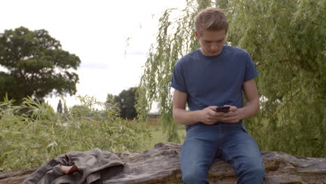 young man sitting on a fallen tree messaging with smartphone