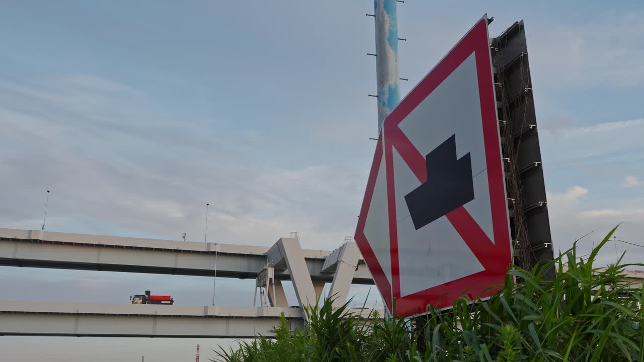 A close-up shot of a prominent junction warning sign with an elevated highway and cloudy sky in the background