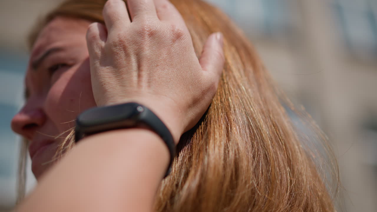 Close up of worried woman adjusting hair under sunlight, wearing black smart watch near blurred building background. Warm tones highlight hair texture and subtle emotion