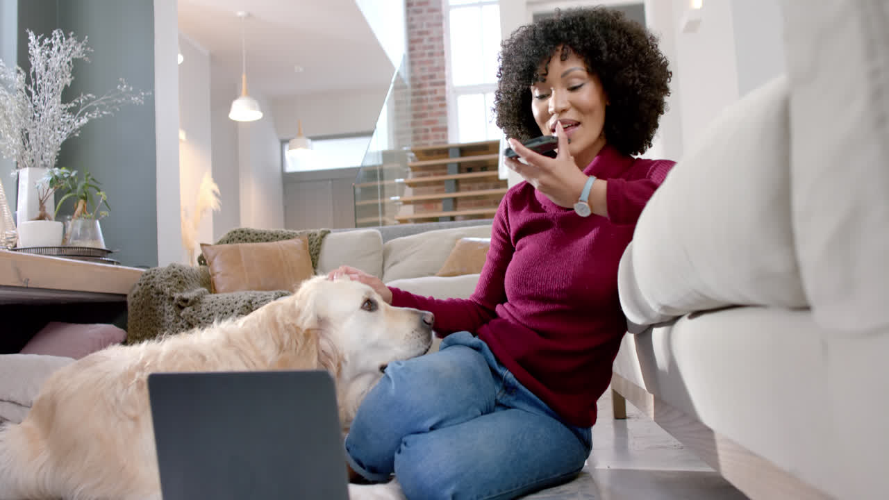 Happy biracial woman with golden retriever dog using laptop and smartphone at home, slow motion