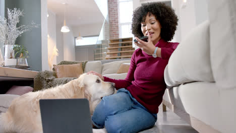 mujer biracial feliz con perro golden retriever usando portátil y teléfono inteligente en casa, cámara lenta