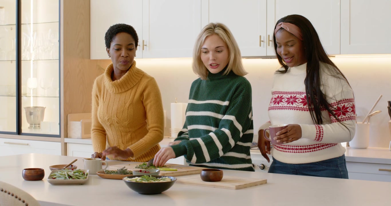 Diverse female friends heeding left woman's pointing, chopping salad into bowls to serve at island