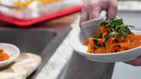chef cleans platting with towel for orange colored pasta noodles infused with tomato paste, carrots or chickpeas in bowls and garnished with fresh parsley sprig, closeup in restaurant or home kitchen