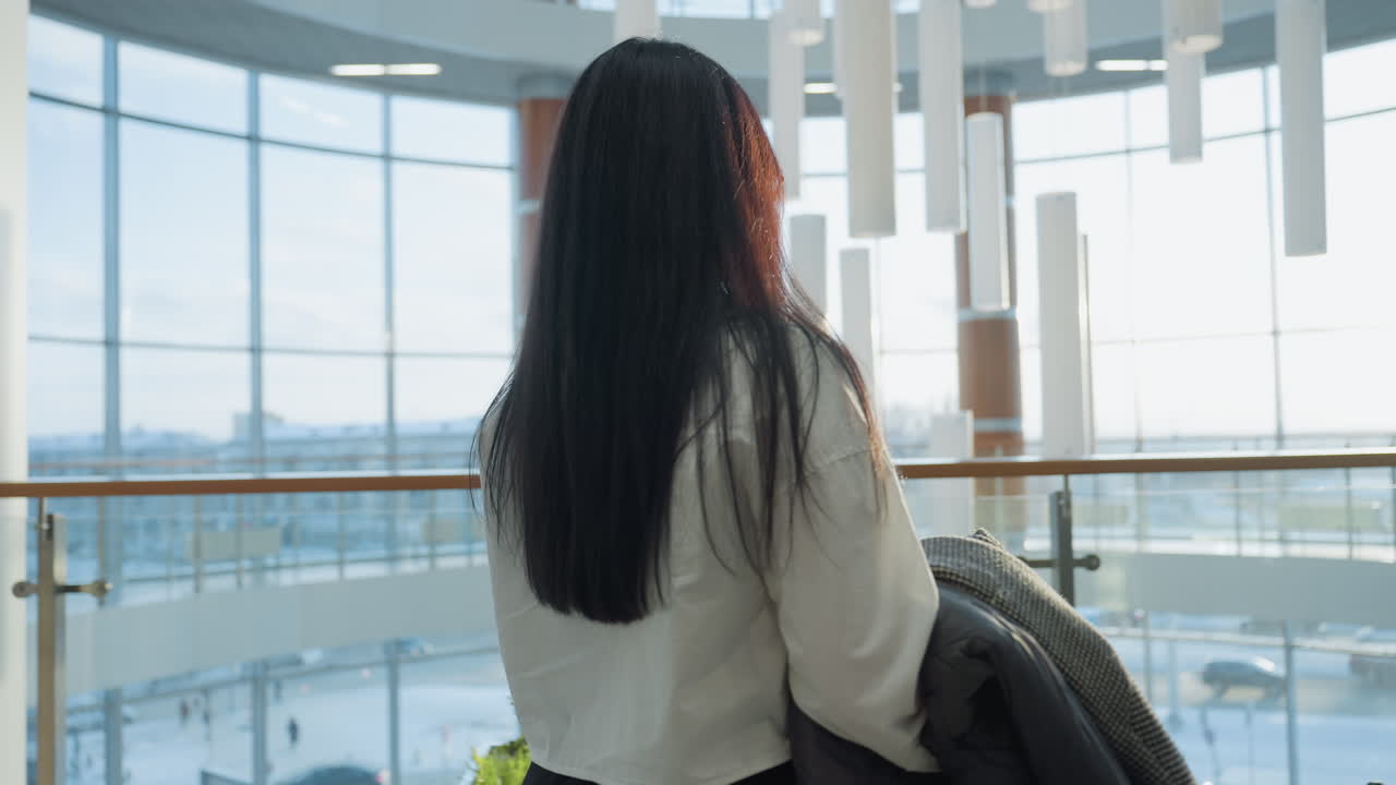 Close up of lady with long hair holding diary and jacket while standing near glass railing inside modern building with sunlight pouring in, sit on nearby decorative bench with green plants
