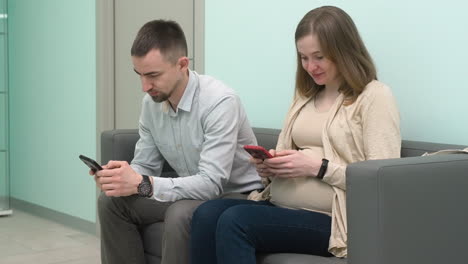 Schwangeres-Paar-Sitzt-Im-Wartezimmer-Und-Schreibt-Auf-Dem-Handy-Eine-SMS