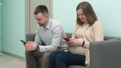 pregnant couple sitting in waiting room and texting on mobile phone