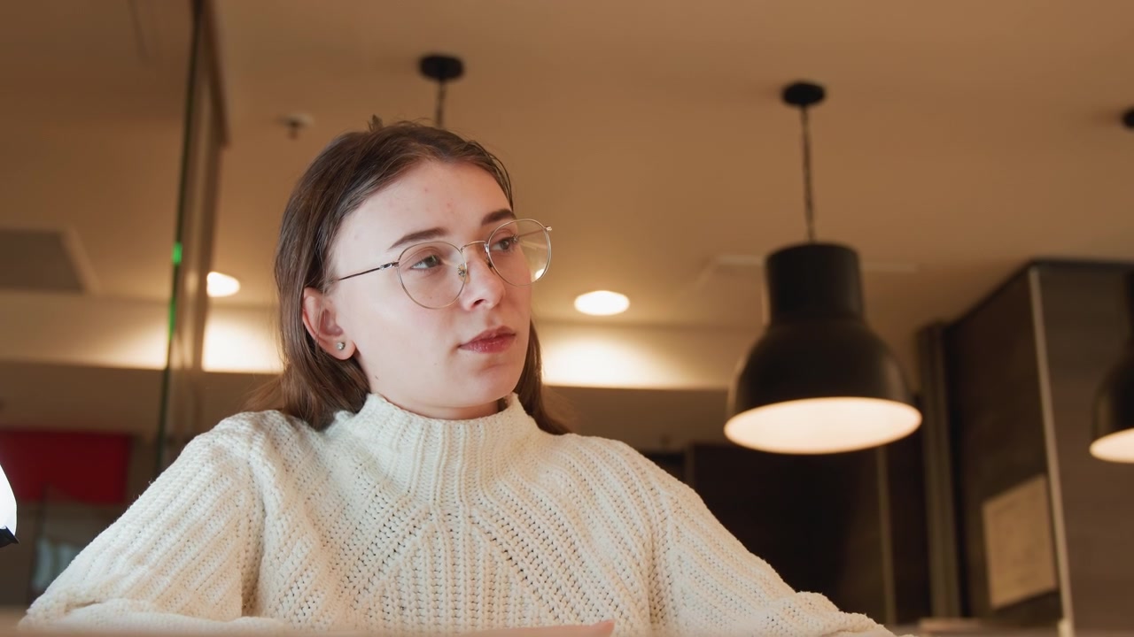 Young woman seated at caf table in warm indoor light holding smartphone, listening with focused gaze and subtle head turn while fingers rest on device, relaxed posture, cozy modern setting