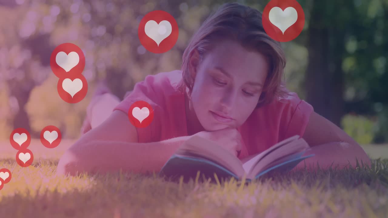animación de iconos de corazón sobre mujer caucásica acostada en el césped y relajándose con un libro