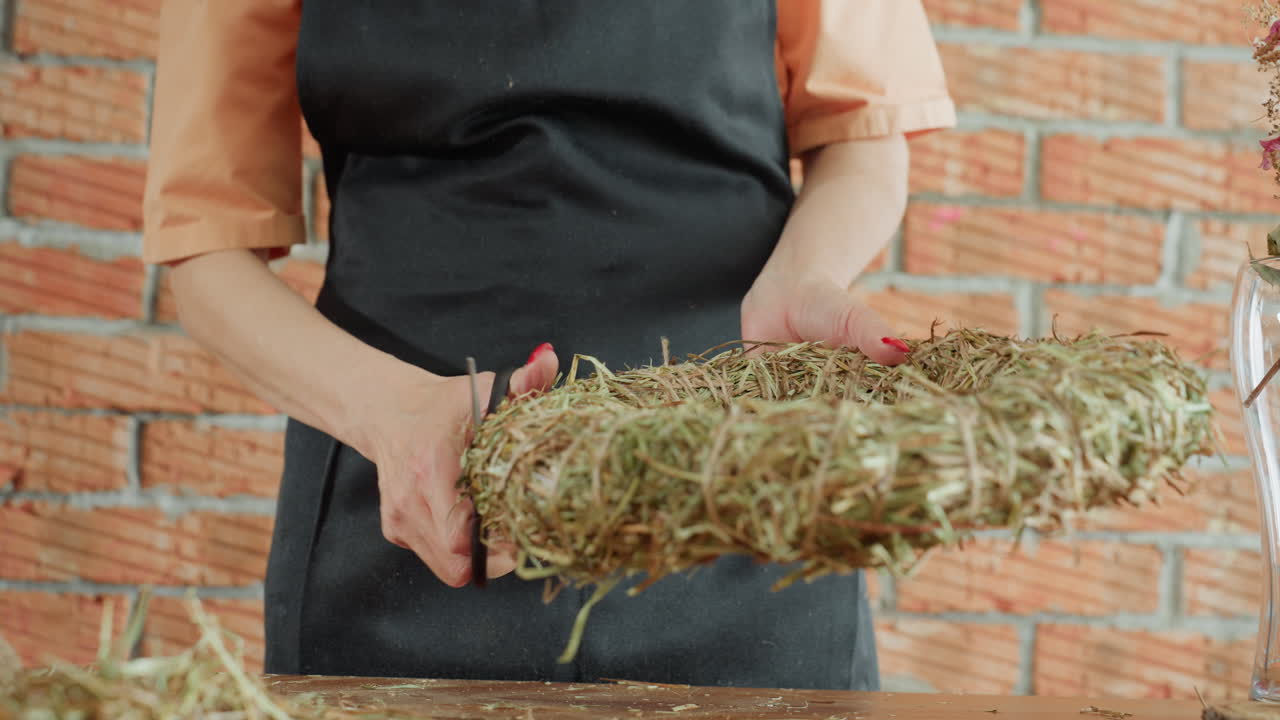 Woman in apron cutting straw wreath with scissors on wooden table, focusing on crafting process, handmade decoration, precision detail, and rustic design in artisan floral arrangement preparation