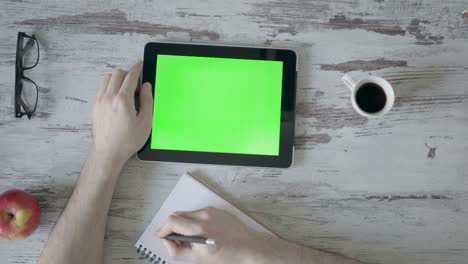 male hands make notes in a notebook, next is a tablet with a green screen