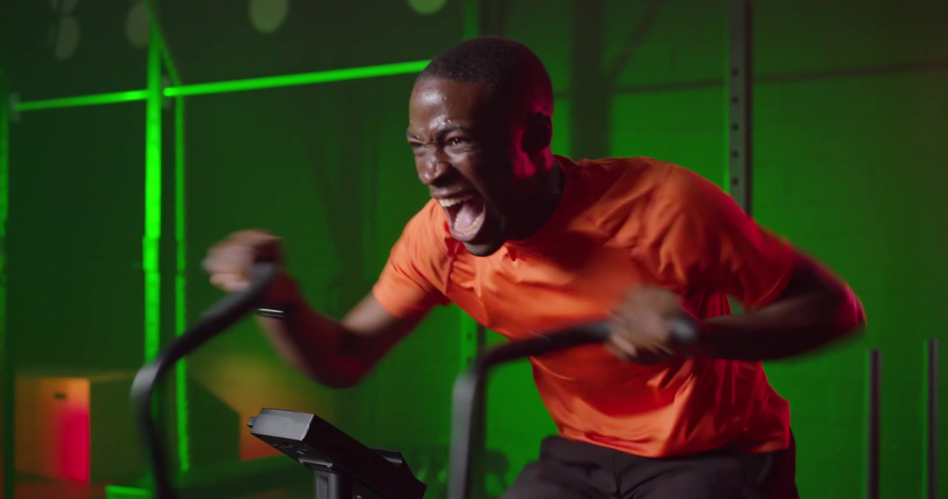 Afroamerikansk mand starter en sprint på en assault bike i et fitnesscenter, cykler i orange trøje for at træne.