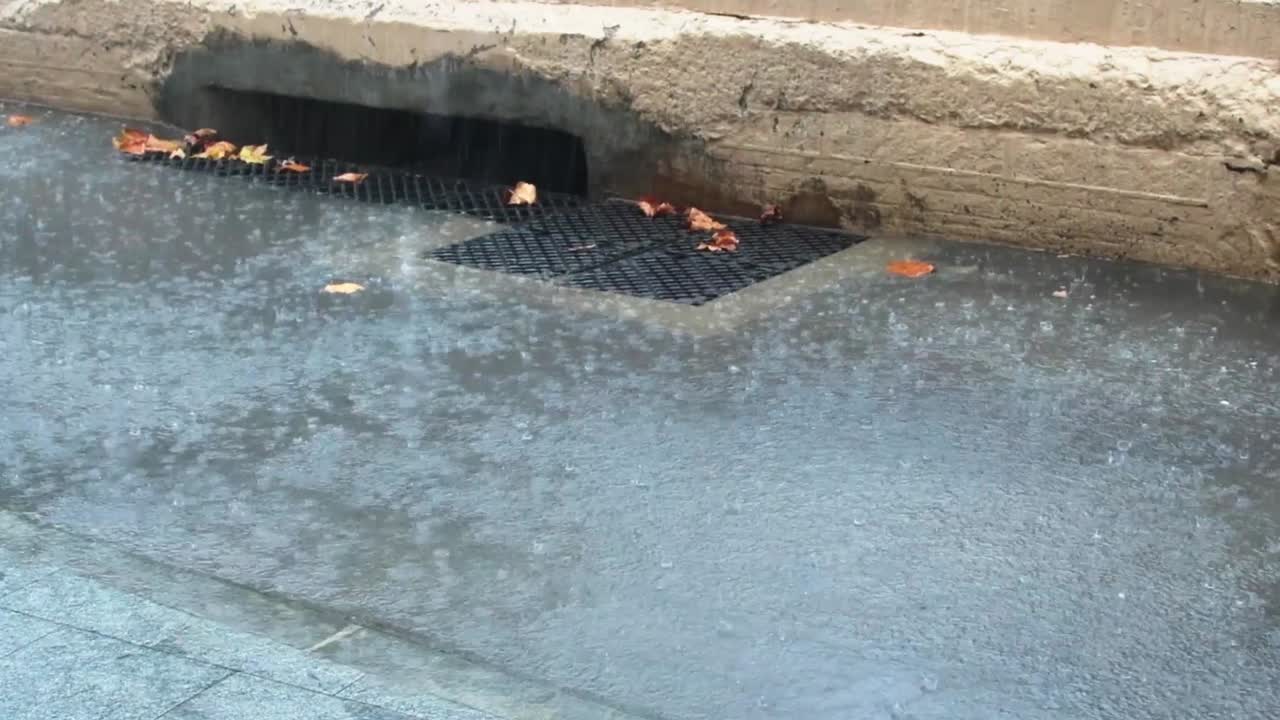 vista de la lluvia cayendo sobre el asfalto cerca de una alcantarilla con hojas en el suelo