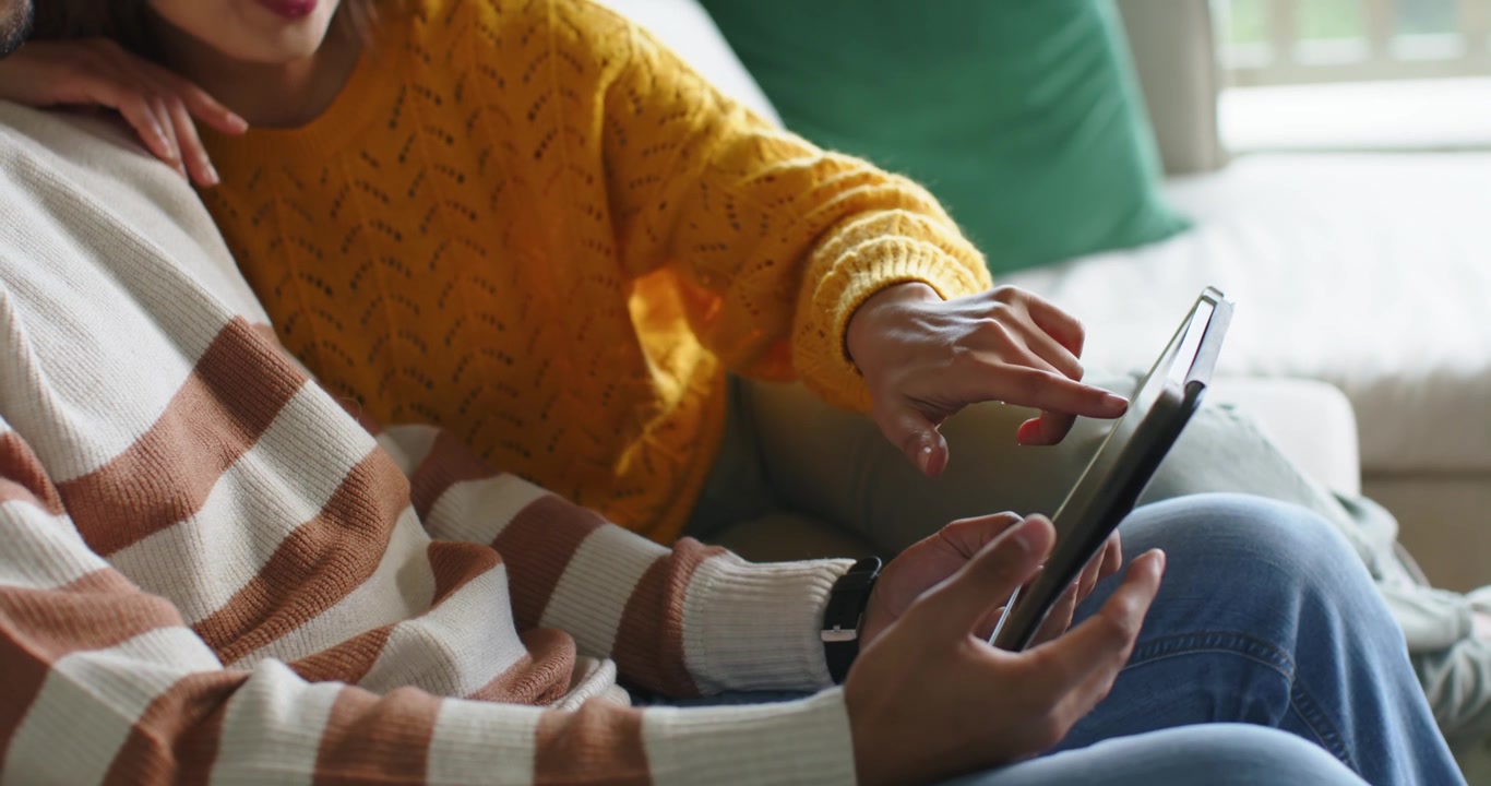 African American couple on couch, woman in mustard sweater tapping tablet to view, man angling it